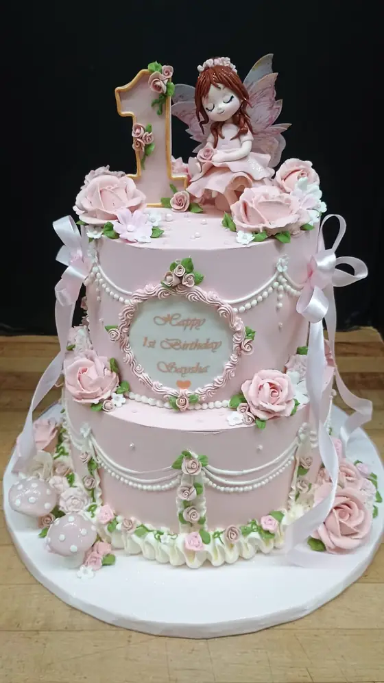 Pink two-tier first birthday cake with fairy topper and buttercream roses